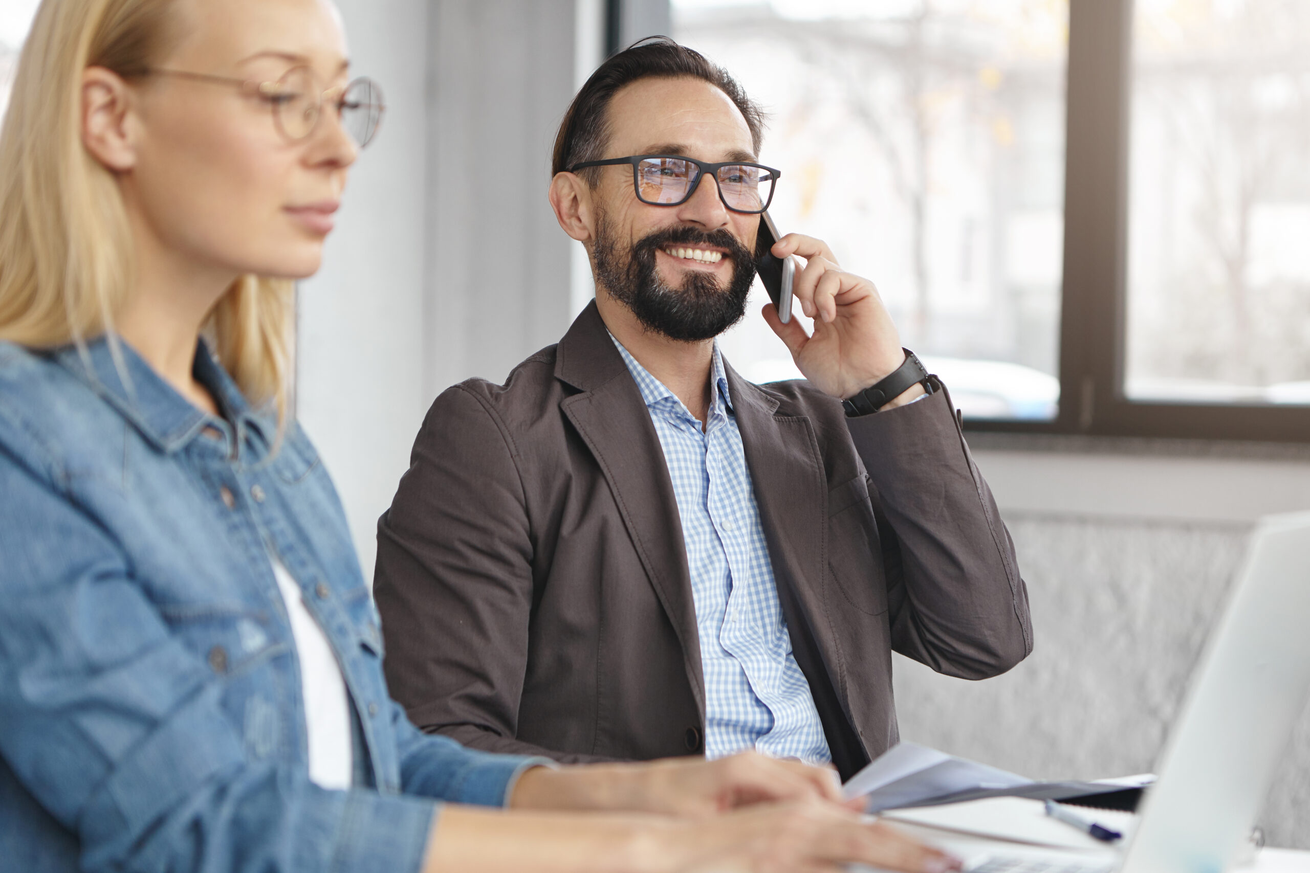 Confident bearded male finacier glad to speak with new investor on smart phone, sits at cabinet near female assistant who creates new project, work together as team. Business coworkers in cabinet